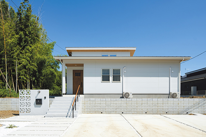 2階建のように見える勾配天井の屋根部分が顔をのぞかせているファサード。真っ白な外壁を玄関ドアや軒天などの木目で引き締め、自然に映える佇まいに。花ブロックをあしらった門柱はリゾート感を演出。